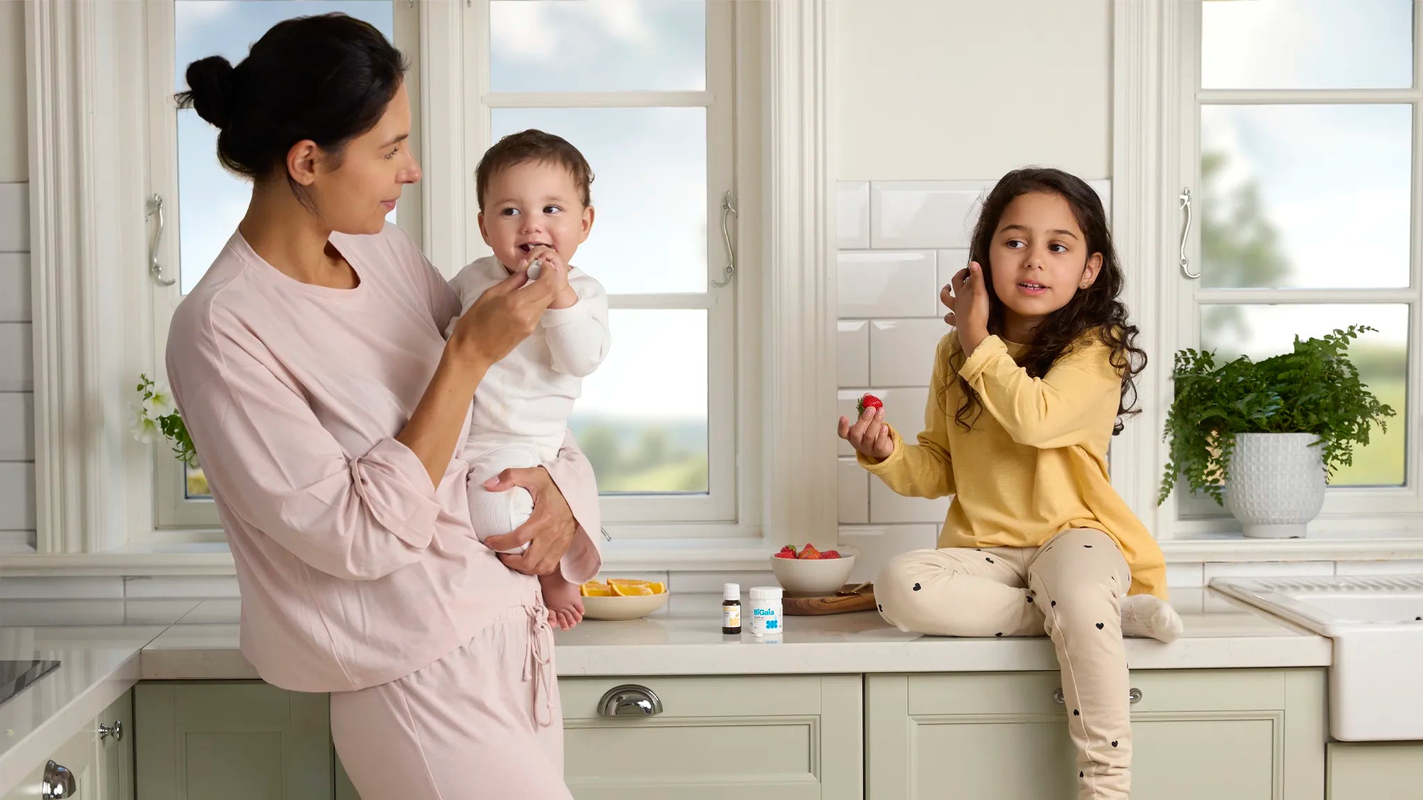 Eine Mutter in der Küche gibt ihrem Kleinkind einen Löffel, während ein älteres Mädchen daneben sitzt; BiGaia Produkte stehen auf der Arbeitsplatte.