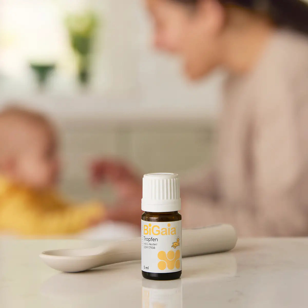 Fokus auf ein Fläschchen BiGaia Tropfen im Vordergrund mit einem unscharfen Baby und Löffel im Hintergrund.