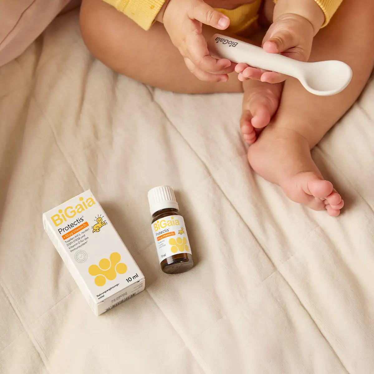BiGaia Protectis Baby-Tropfen Packung und Fläschchen auf einer Decke neben den Füßen eines Babys, das einen Löffel hält.