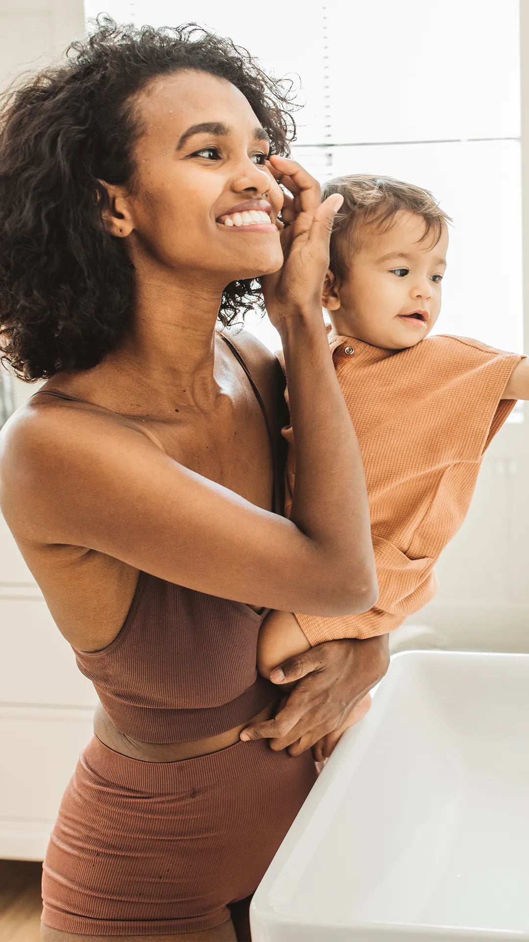Eine Frau hält ein Kleinkind im Arm vor einem Badezimmerspiegel; beide schauen lächelnd hinein, was tägliche Pflegeroutinen symbolisiert.