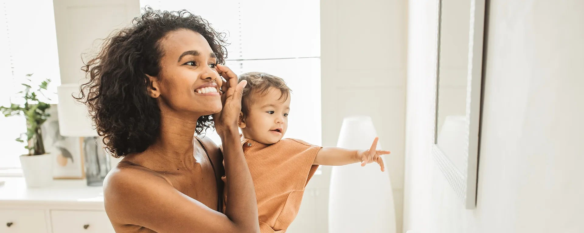 Eine Frau hält ein Kleinkind im Arm vor einem Badezimmerspiegel; beide schauen lächelnd hinein, was tägliche Pflegeroutinen symbolisiert.