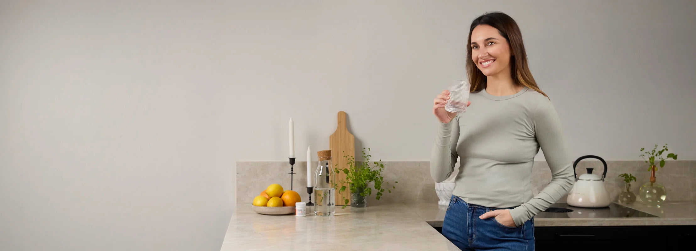 Eine lächelnde Frau steht in einer modernen Küche und hält ein Glas Wasser bereit, neben ihr steht eine Packung BiGaia Kapseln.