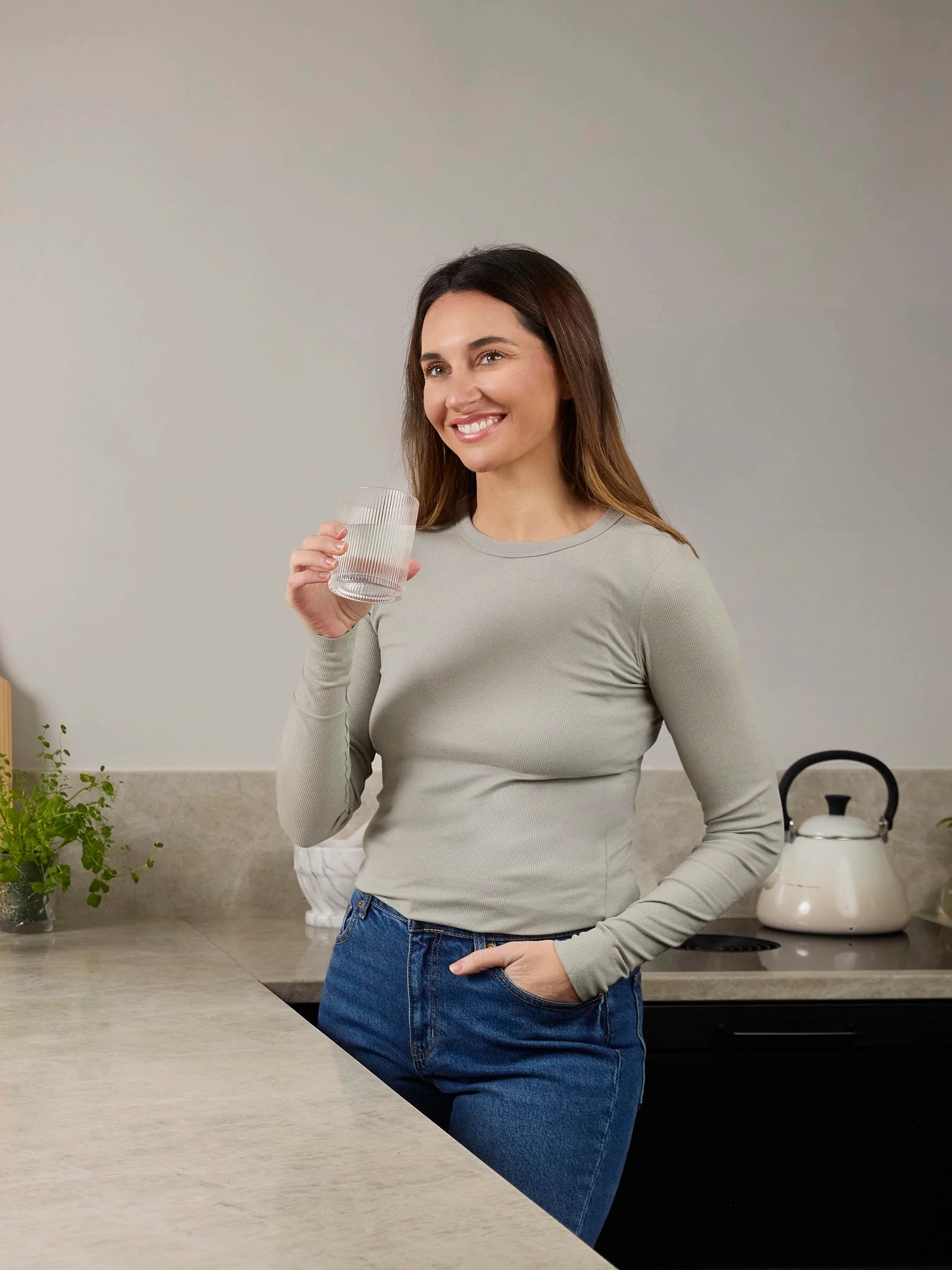 Eine lächelnde Frau steht in einer modernen Küche und hält ein Glas Wasser bereit, neben ihr steht eine Packung BiGaia Kapseln.