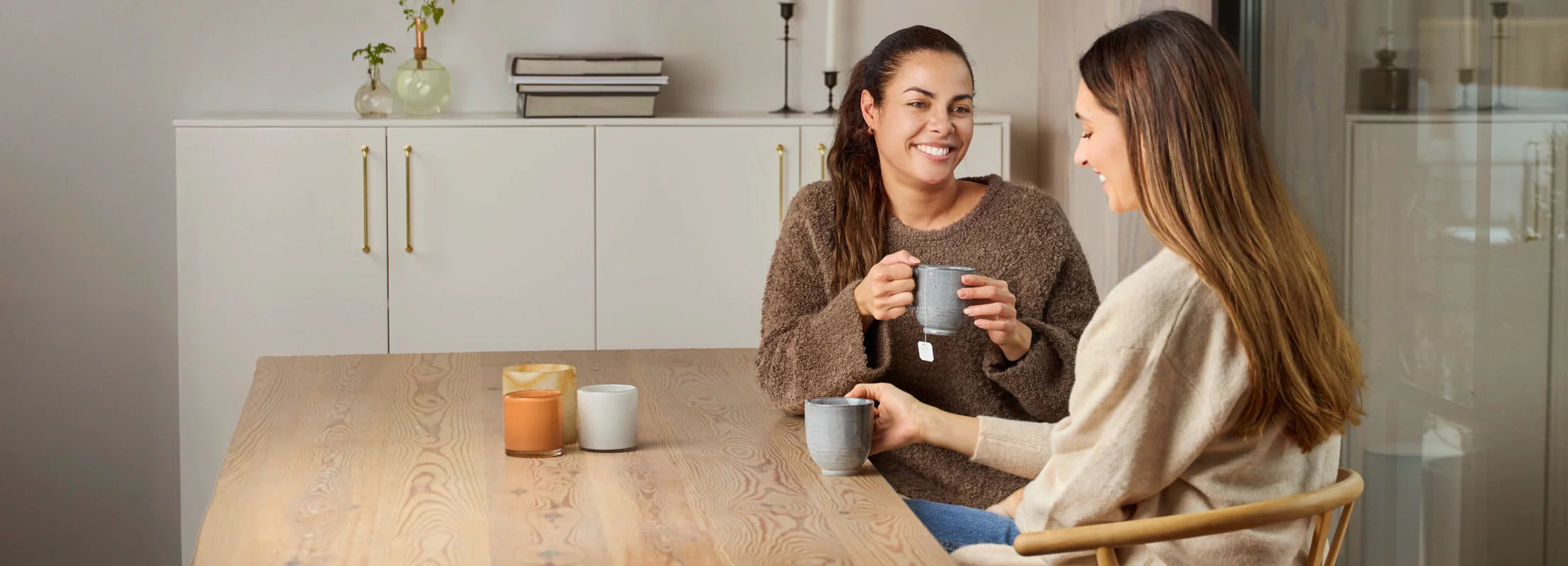 Zwei Frauen sitzen entspannt an einem Holztisch, trinken Tee und unterhalten sich lächelnd in einem hellen Wohnraum.