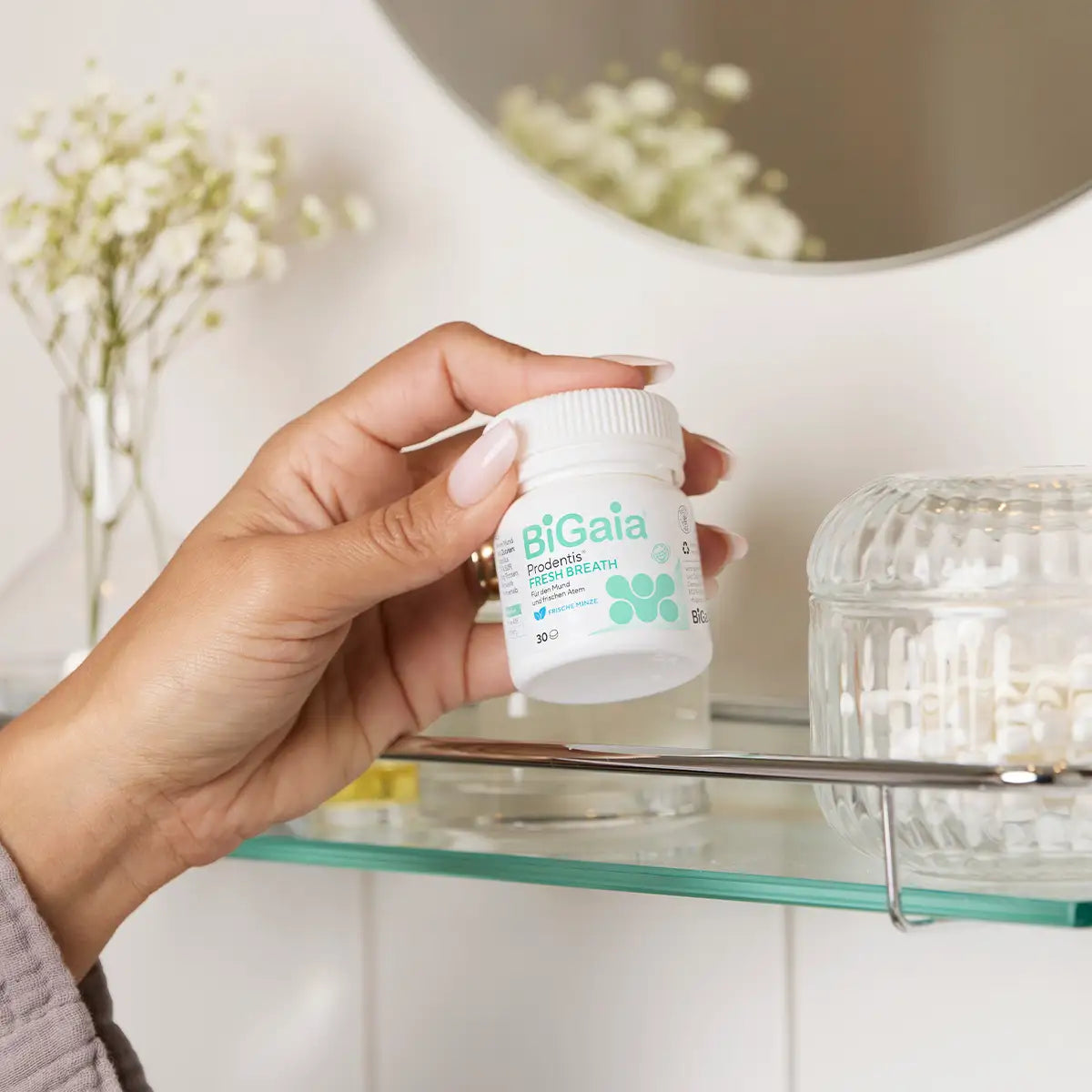 Eine Hand nimmt die Dose BiGaia Prodentis Fresh Breath von einem gläsernen Badezimmerregal neben einem Spiegel und weißen Blumen.