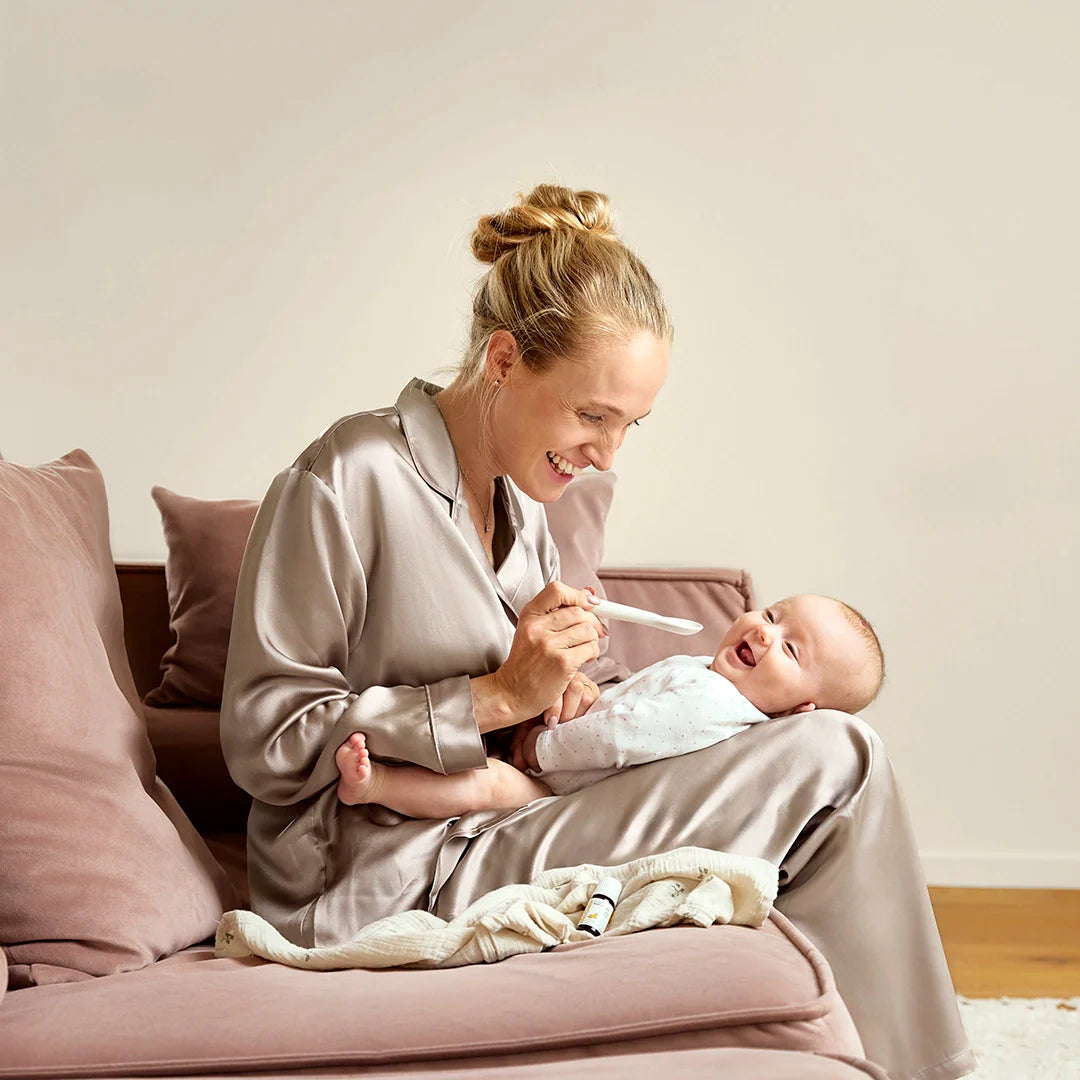 Eine lachende Mutter sitzt auf einem Sofa und gibt ihrem fröhlichen Baby, das auf ihrem Schoß liegt, BiGaia-Tropfen per Löffel.